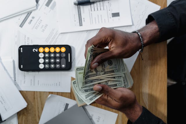 pexels-tima-miroshnichenko-6694570 psomeone counting cash at a table covered in papers with a calculator open
