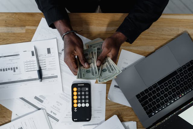 person-sitting-at-desk-counting-money-and-using-a-calculator-with-papers-and-a-laptop-around-them person-sitting-at-desk-counting-money-and-using-a-calculator-with-papers-and-a-laptop-around-them